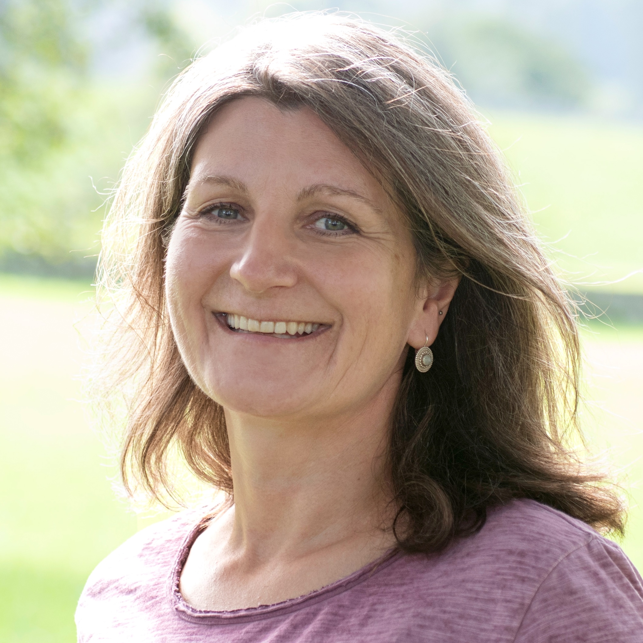 Lächelnde Ursula Hess  mit braunen Haaren, die vor einem grünen, unscharfen Hintergrund steht.