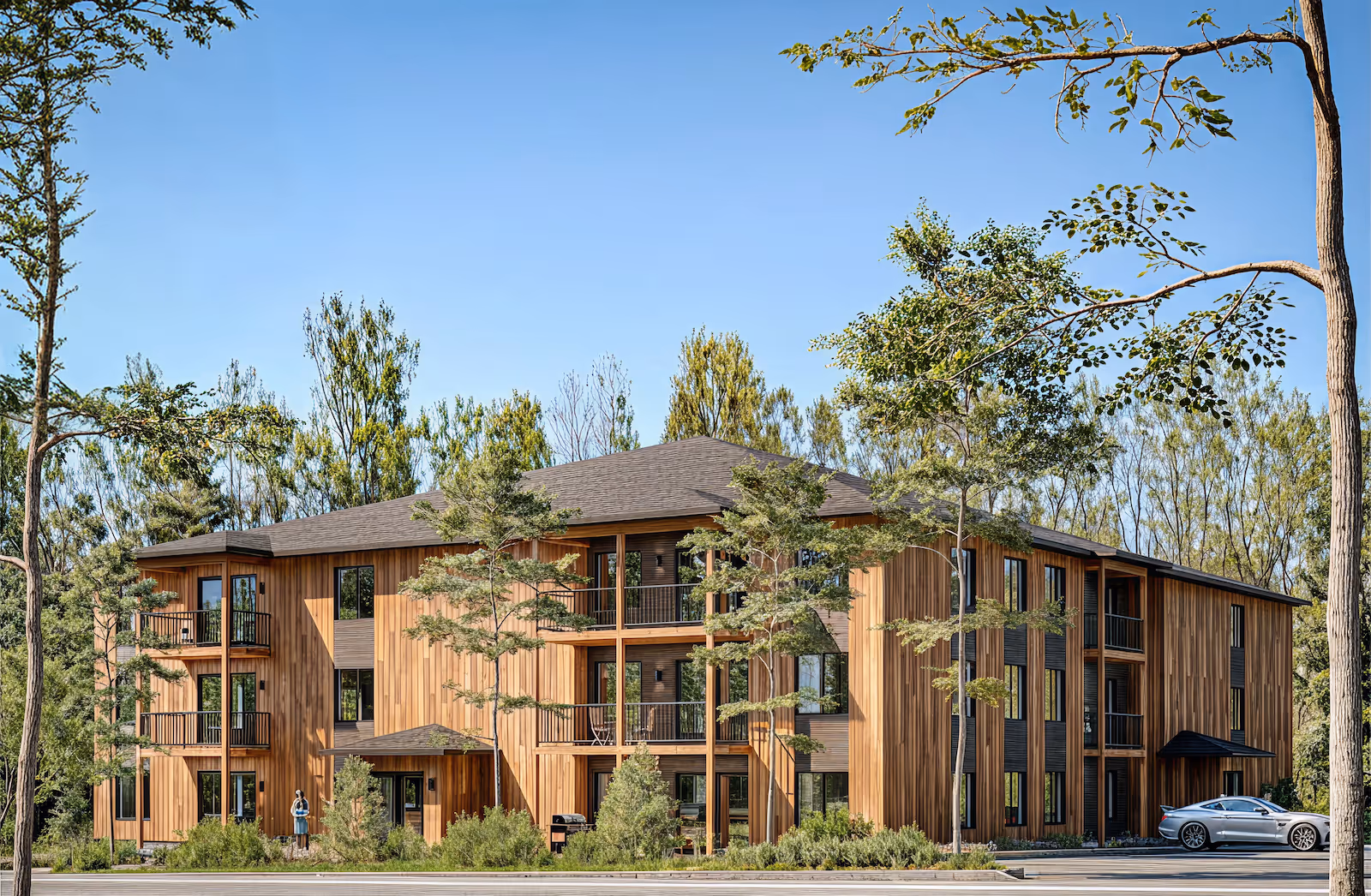 Immeuble résidentiel moderne en bois sur trois étages entouré d’arbres avec une voiture grise garée à droite.