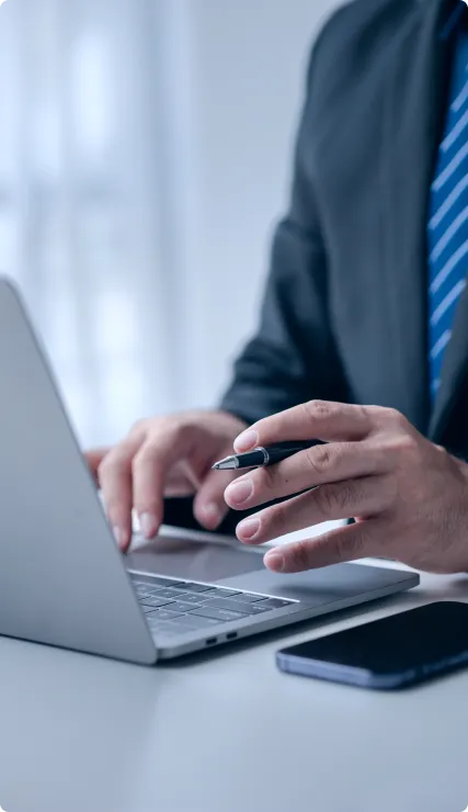 Person in suit typing on laptop with pen in hand and smartphone on white desk.