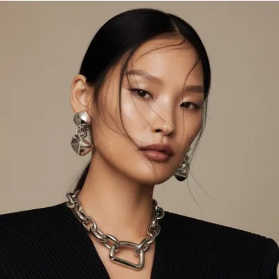 Portrait of a woman with sleek black hair, wearing large silver earrings and a chunky silver chain necklace with a black top.