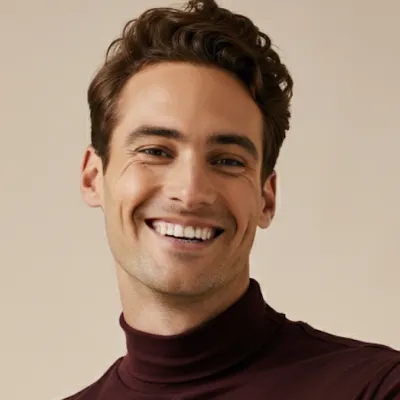 Smiling man with short curly brown hair wearing a dark maroon turtleneck against a beige background.