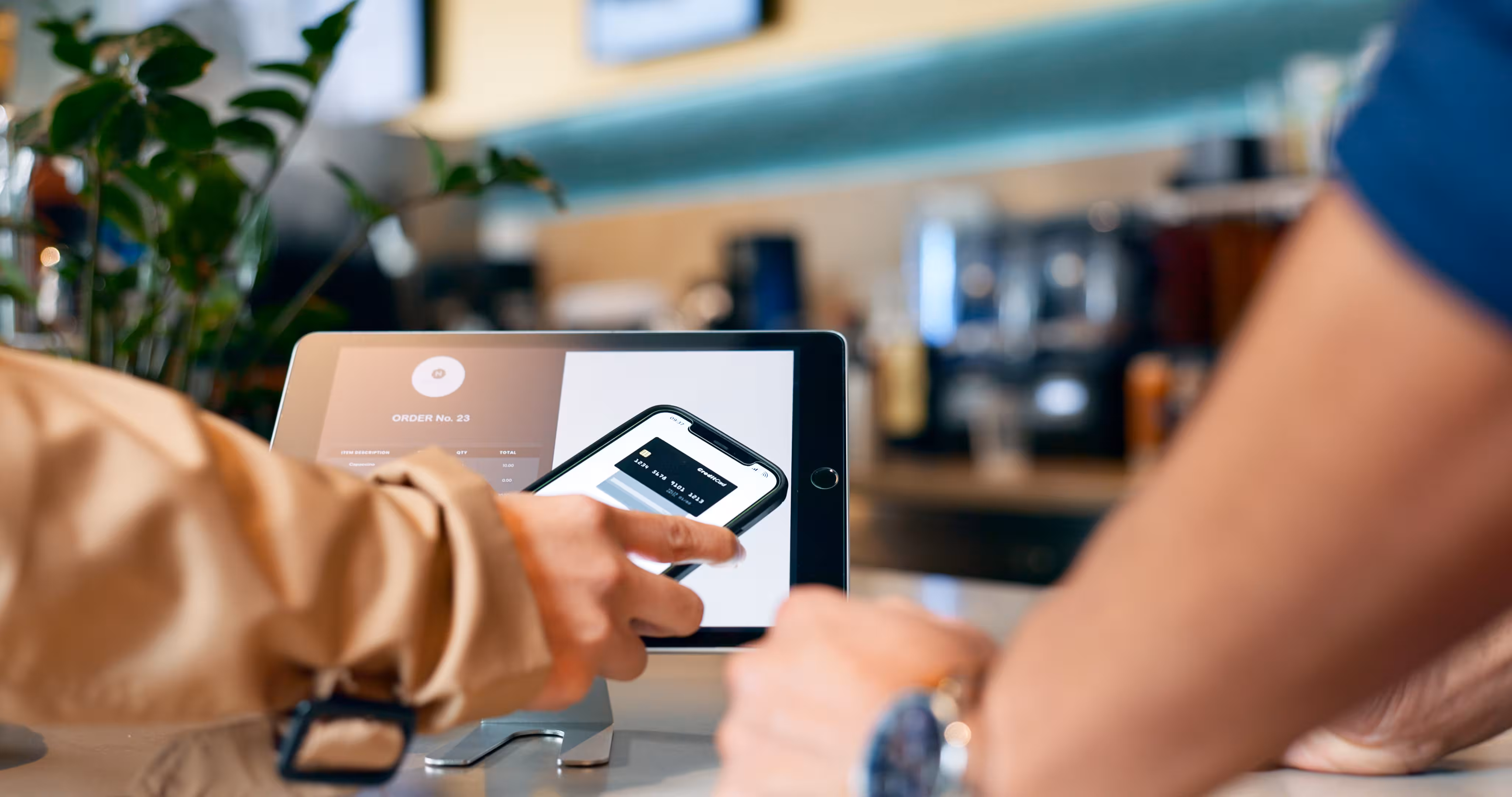 Person using a smartphone to complete a digital payment on a tablet screen in a café setting.