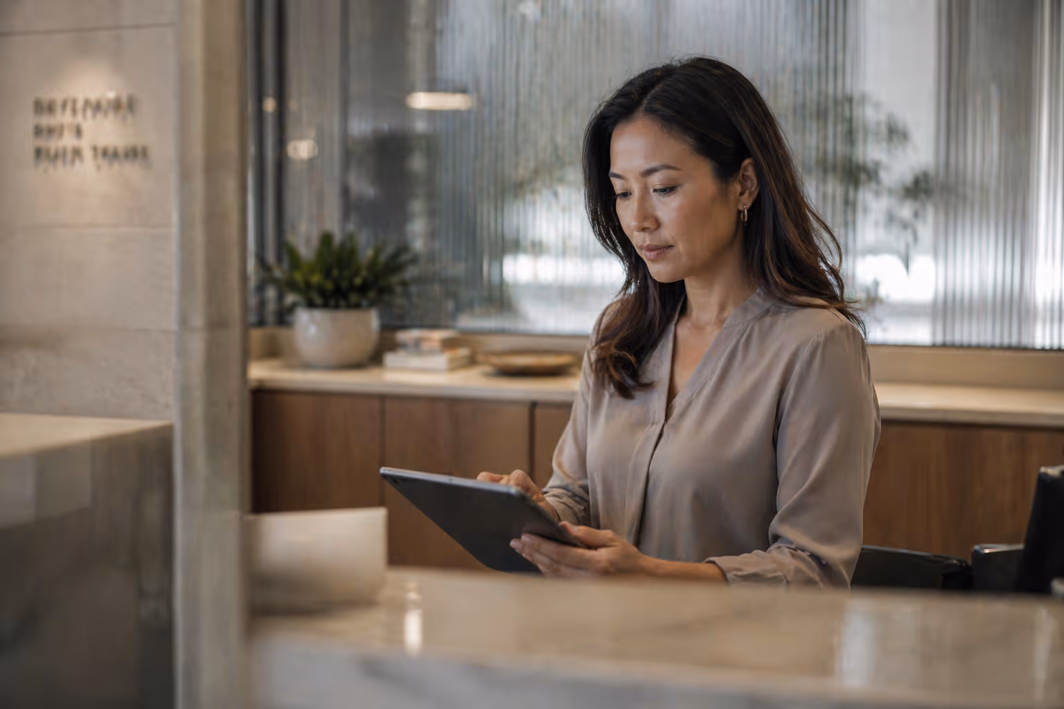 Woman sitting at a wooden table using a tablet in a modern office space with plants and blurred background.