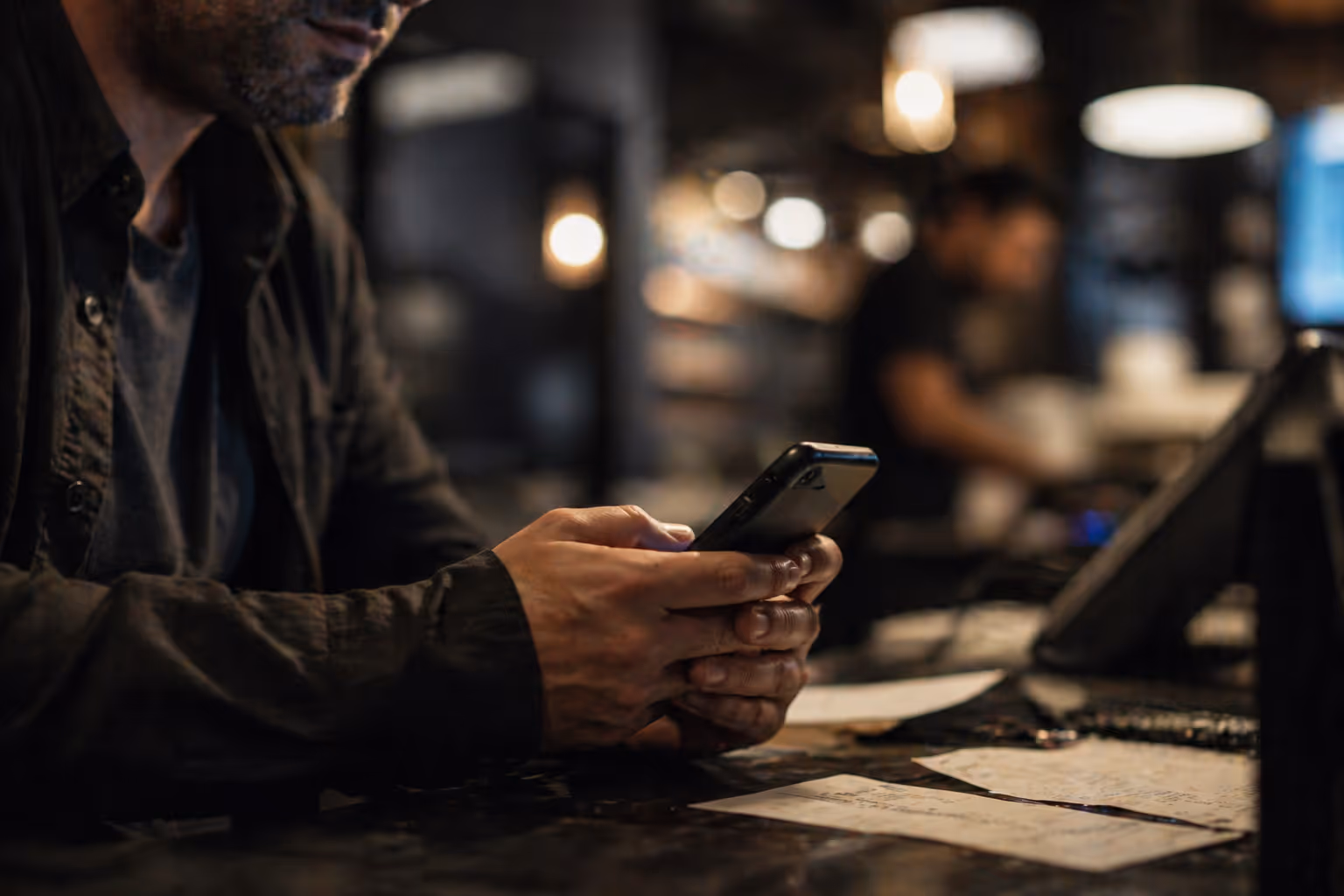 Person wearing a dark jacket using a smartphone at a dimly lit countertop with scattered papers.