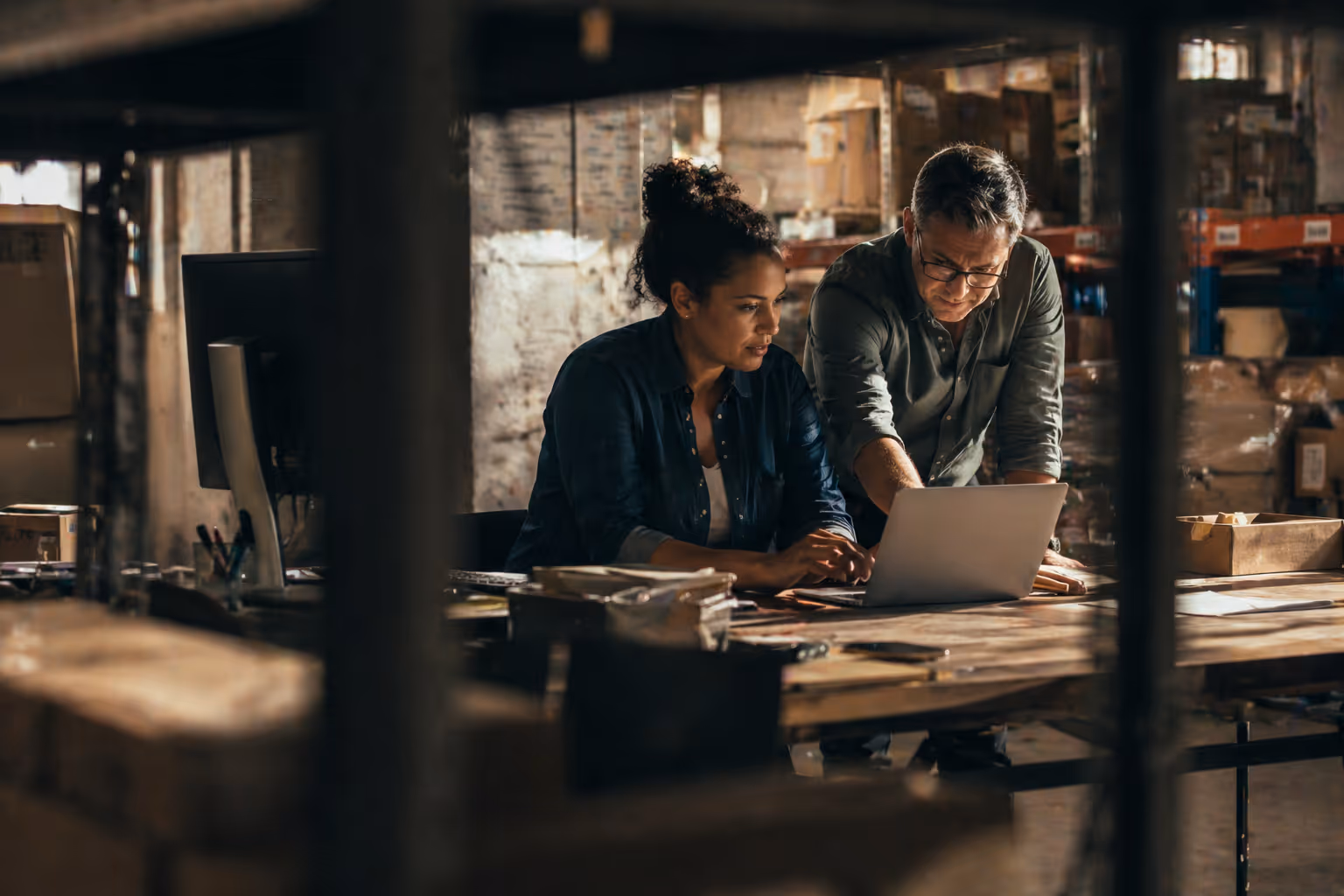 Two coworkers collaborating on a laptop in a warehouse office filled with cardboard boxes.
