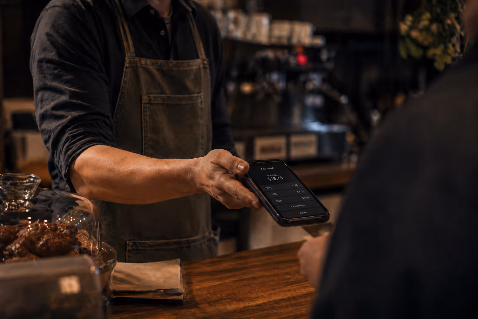 Barista holding a smartphone showing a bill of $18.75 with tip options at a coffee shop counter.