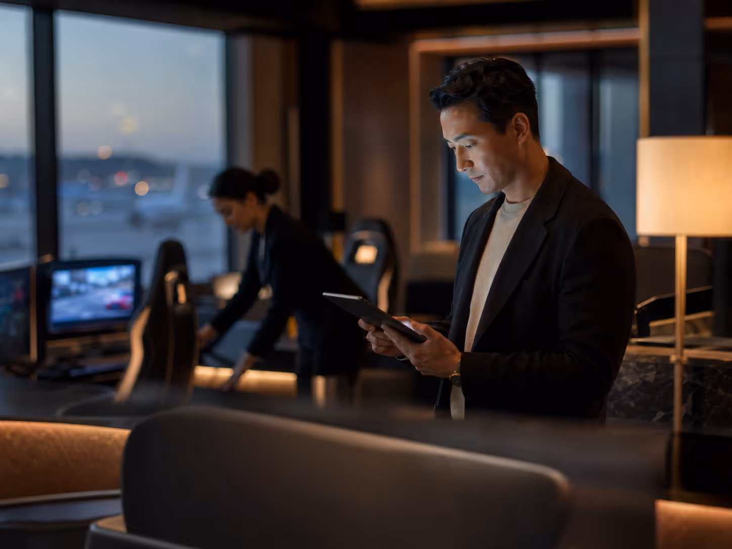 Man in blazer using a tablet in a dimly lit modern office with a woman working near a computer in the background.