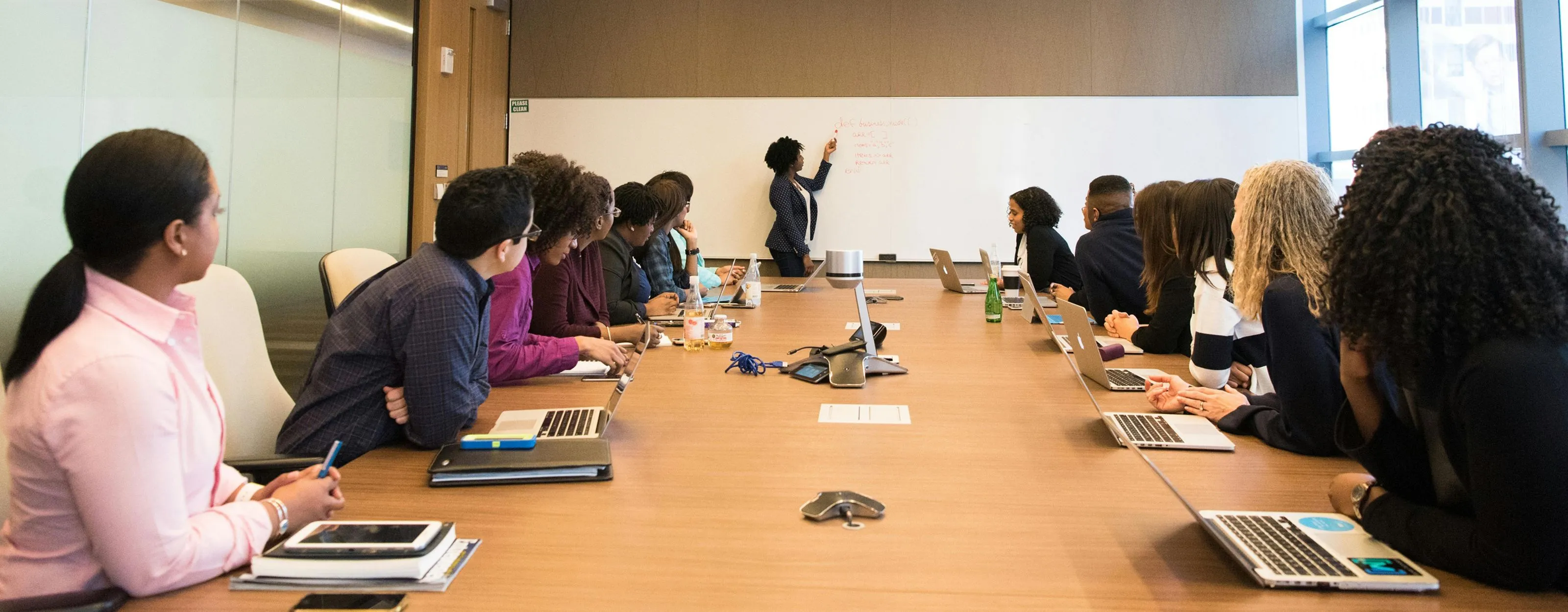 Diverse group of professionals seated around a conference table with laptops, listening to a woman presenting at a whiteboard in a modern office.