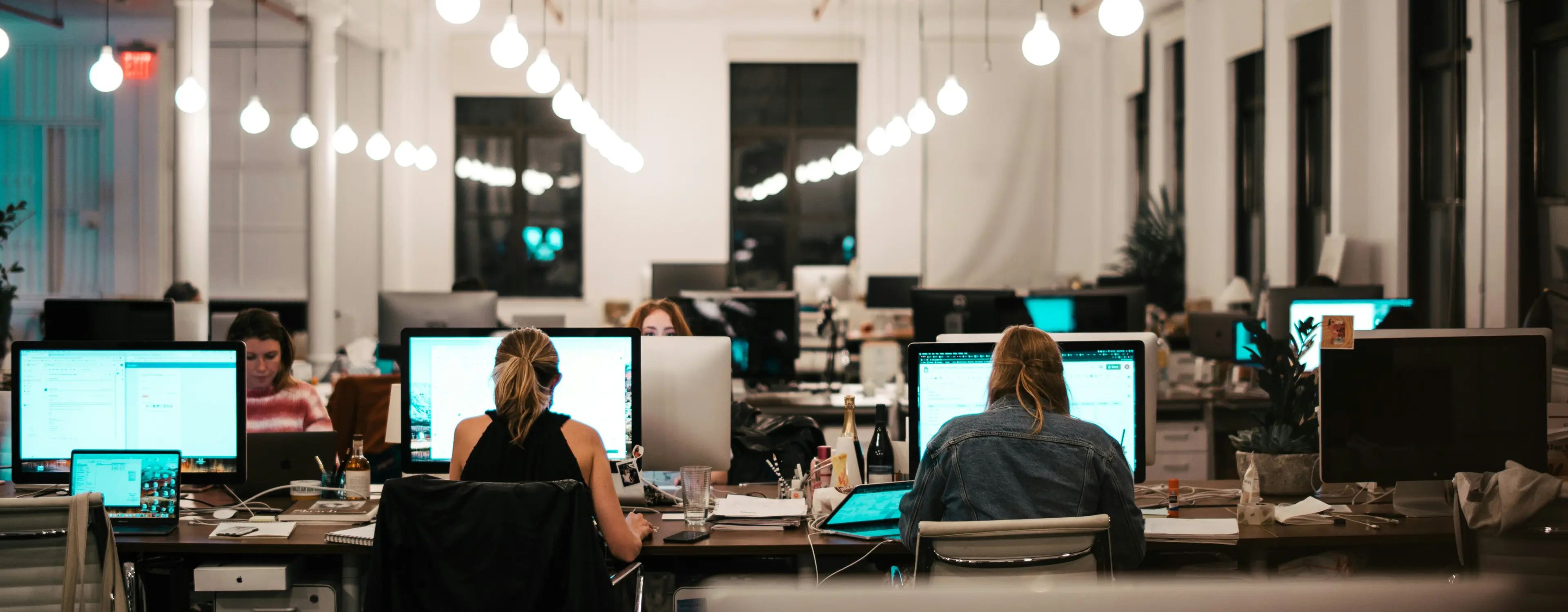 Open office workspace with people working at desks on computers under hanging round light bulbs.