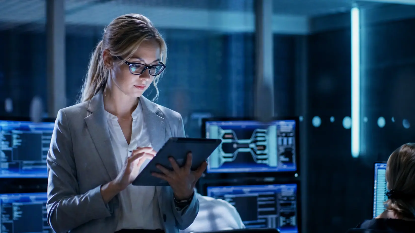 Woman in glasses and a blazer using a tablet in a high-tech control room with multiple monitors displaying data and diagrams.