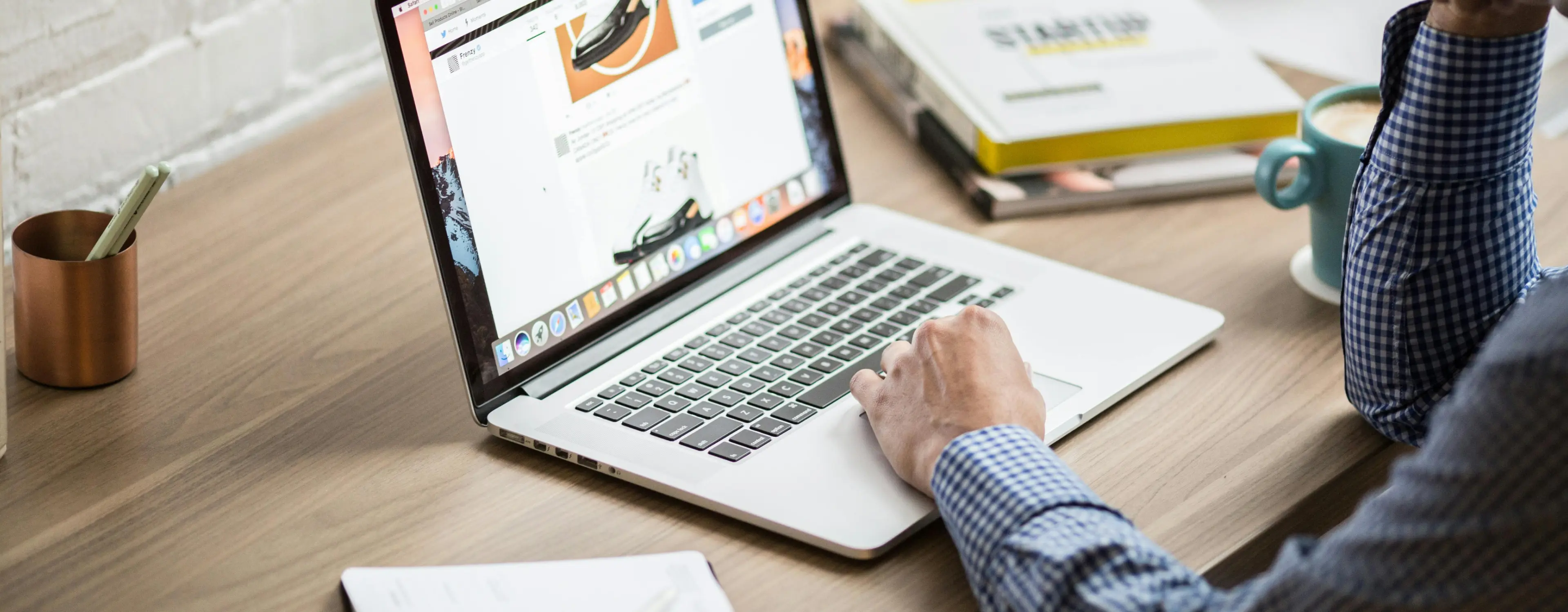Person in a blue checkered shirt using a laptop on a wooden desk with a cup of coffee and books nearby.
