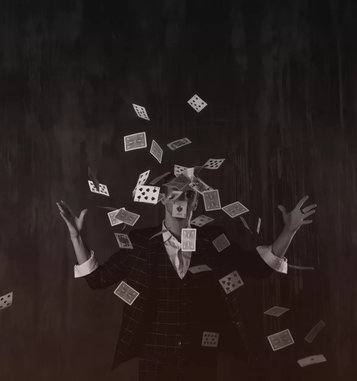 Man in checkered suit tossing playing cards in the air against a dark background.