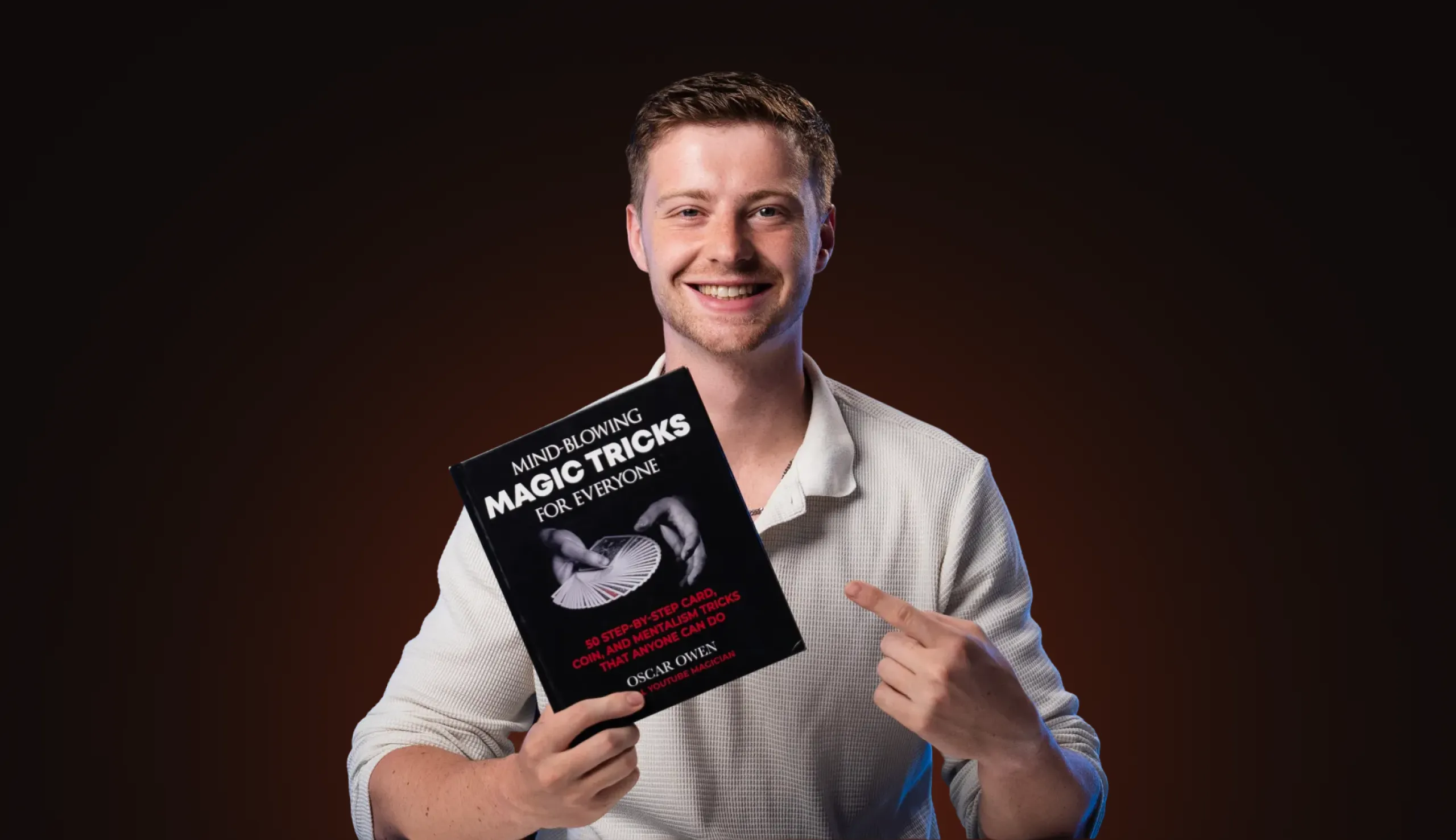 Smiling man in white shirt holding and pointing to a book titled 'Mind-Blowing Magic Tricks for Everyone' against a dark background.