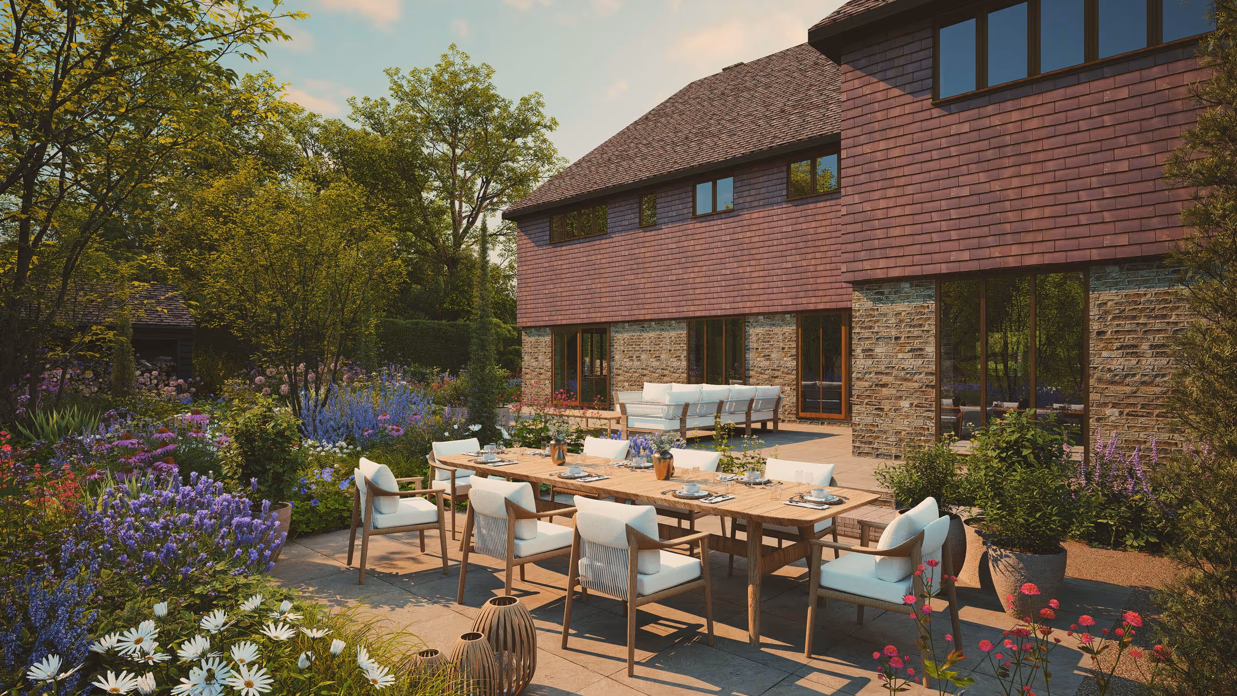 Spacious outdoor patio with a long wooden dining table set with white cushioned chairs, surrounded by lush flowering plants and a modern stone and shingle house.