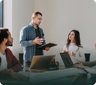 A man standing and explaining something to colleagues seated around a table with laptops in a bright office.