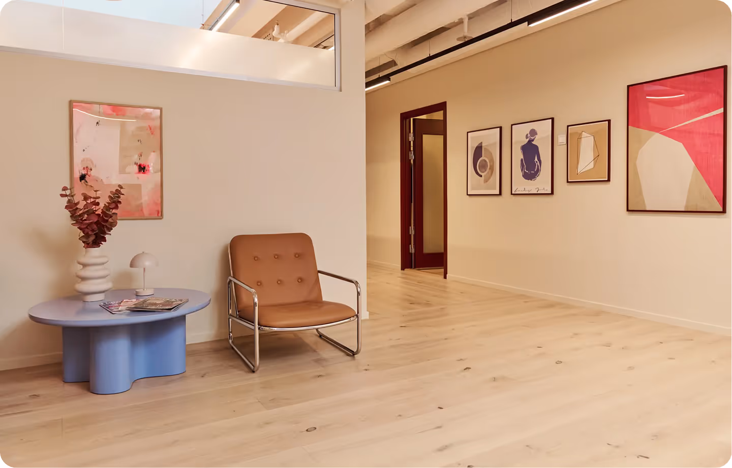 Väntrum med ljusblått runt bord, brunt läderfåtölj, kruka med röd växt och abstrakta konstverk på väggarna.