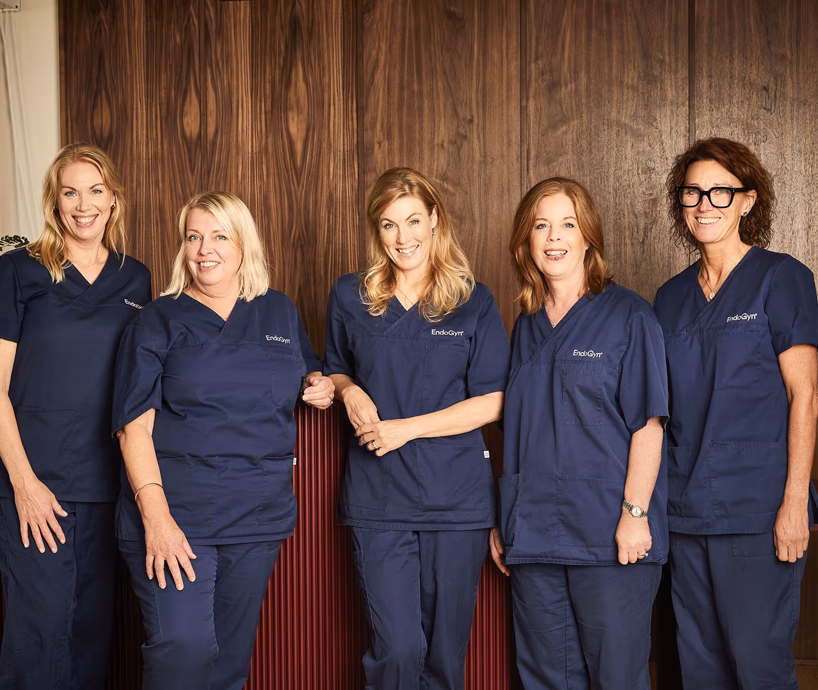 Fem kvinnor i blå medicinska uniformer står leende framför en mörk träpanelvägg.