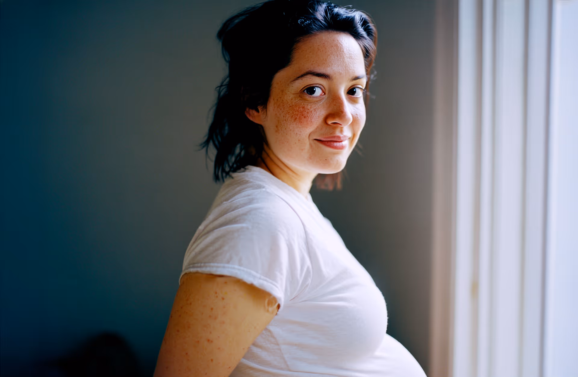 Gravid kvinna med vit t-shirt som ler och ser mot kameran medan hon står bredvid ett fönster.