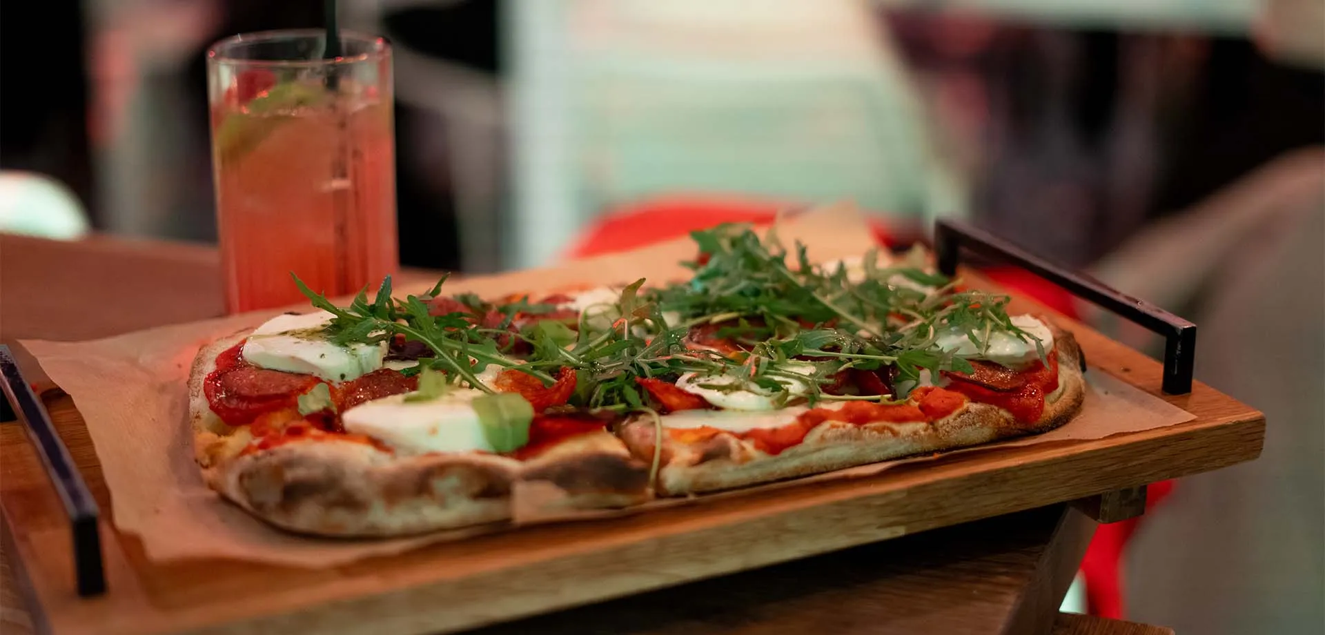 Rechteckige Pizza mit Pepperonie, frischer Mozzarella, auf einem Holz-Servierbrett neben einem Getränk.