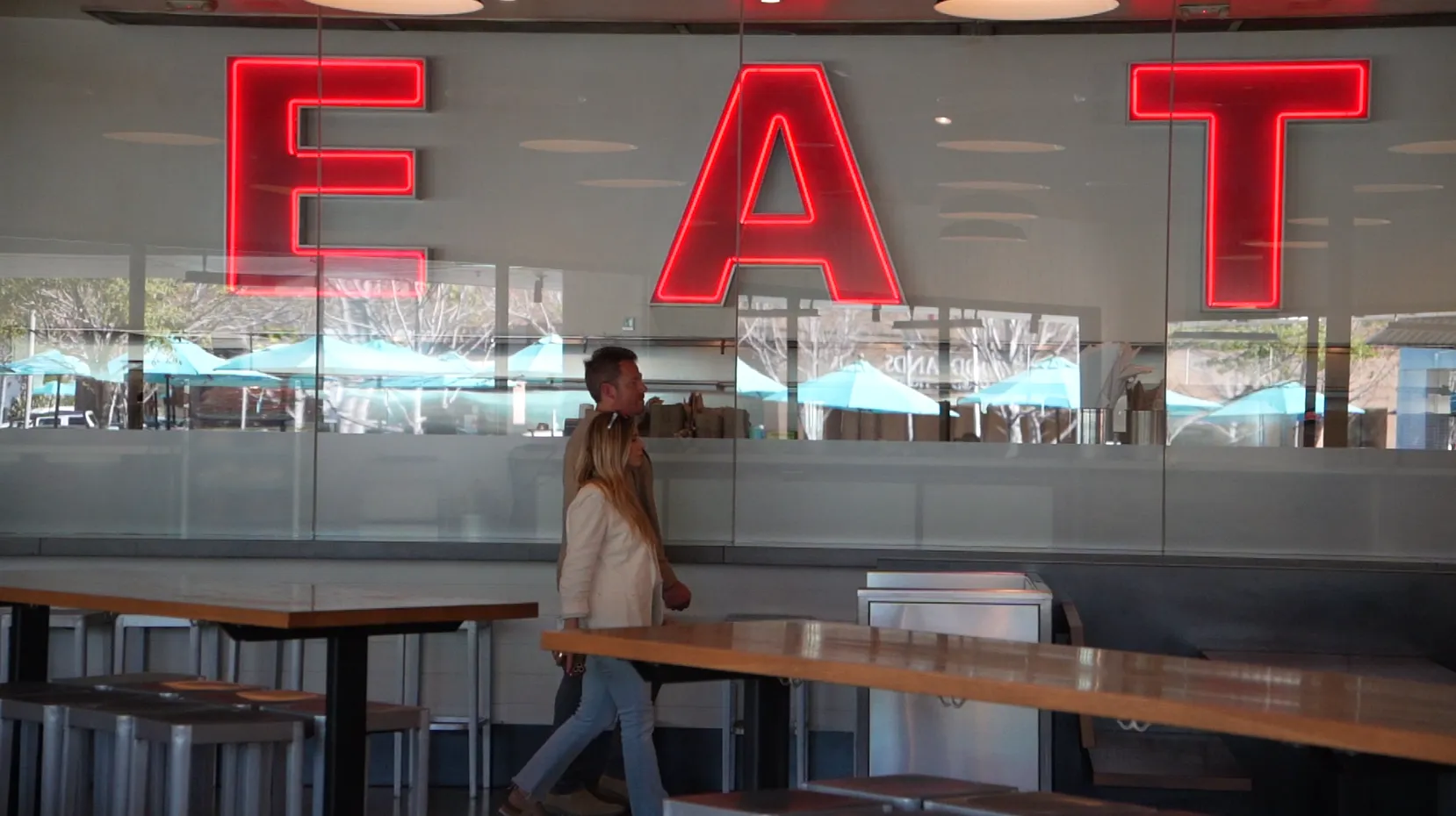 Two people walking inside a restaurant with large red neon letters spelling 'EAT' on the glass wall behind them.