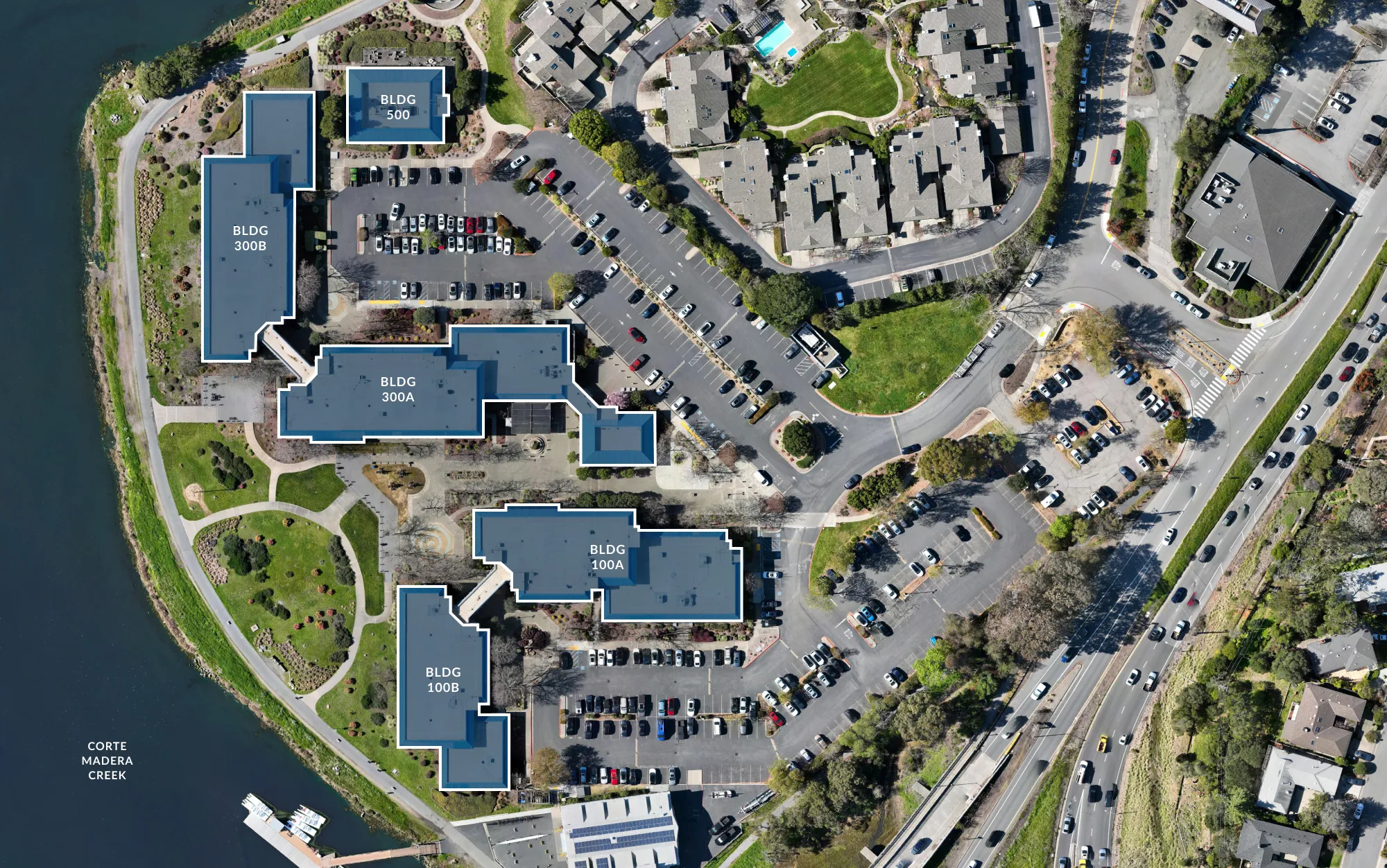 Aerial view of a property complex featuring labeled buildings 100A, 100B, 300A, 300B, and 500 surrounded by parking lots, green areas, walkways, residential houses, and a water body named Corte Madera Creek.
