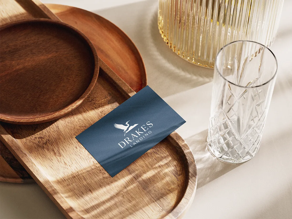 A navy blue business card with a white logo and the text 'DRAKES LANDING' placed on a wooden tray, next to an empty clear textured glass and a decorative glass item on a light-colored surface.