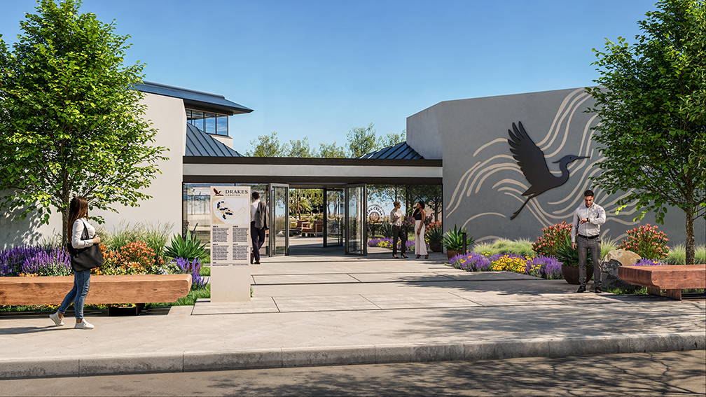 Exterior view of a modern courtyard entrance with glass doors, landscaped with trees and colorful flowers, wooden benches on each side, and a large metal bird sculpture mounted on a beige wall, with people walking and standing nearby under a clear blue sky.