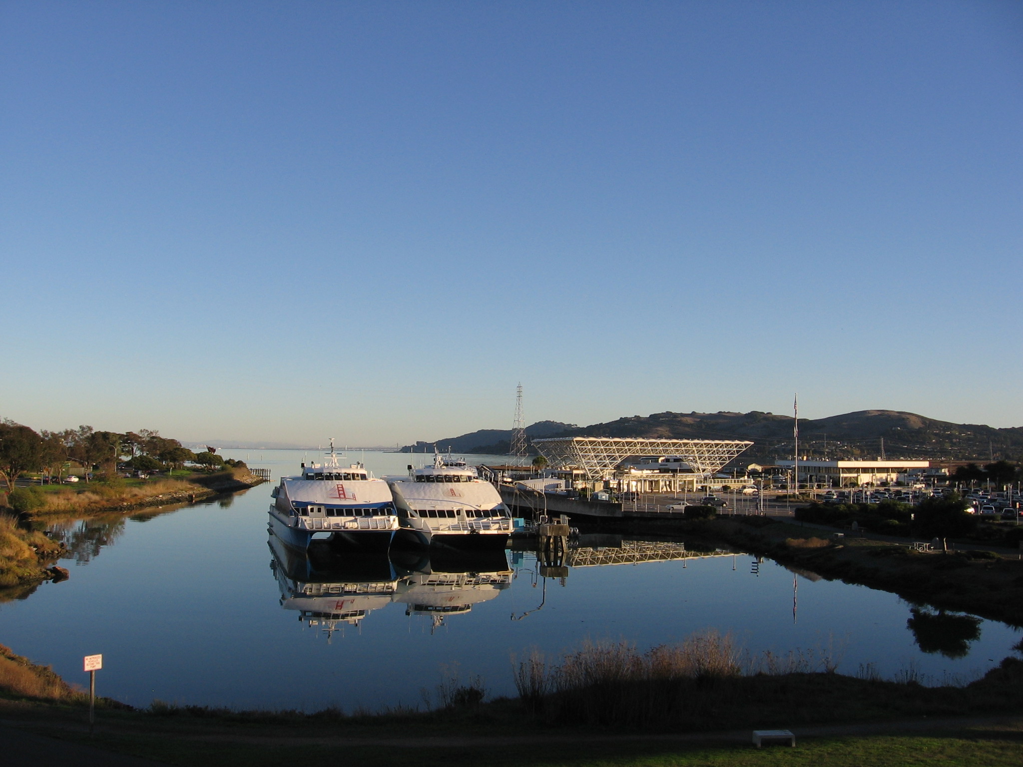 Two white ferries docked side by side on calm water reflecting the vessels and nearby structures, with hills and a clear blue sky in the background.