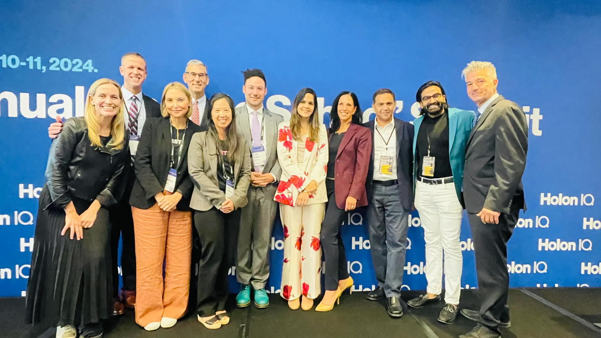Group of eleven smiling professionals standing on stage at Holon IQ event with blue backdrop.