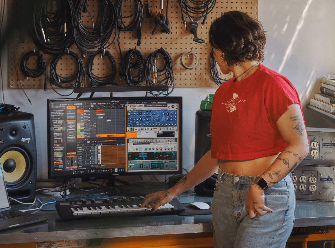 Person in red shirt using a MIDI keyboard connected to a computer with music software on screen.