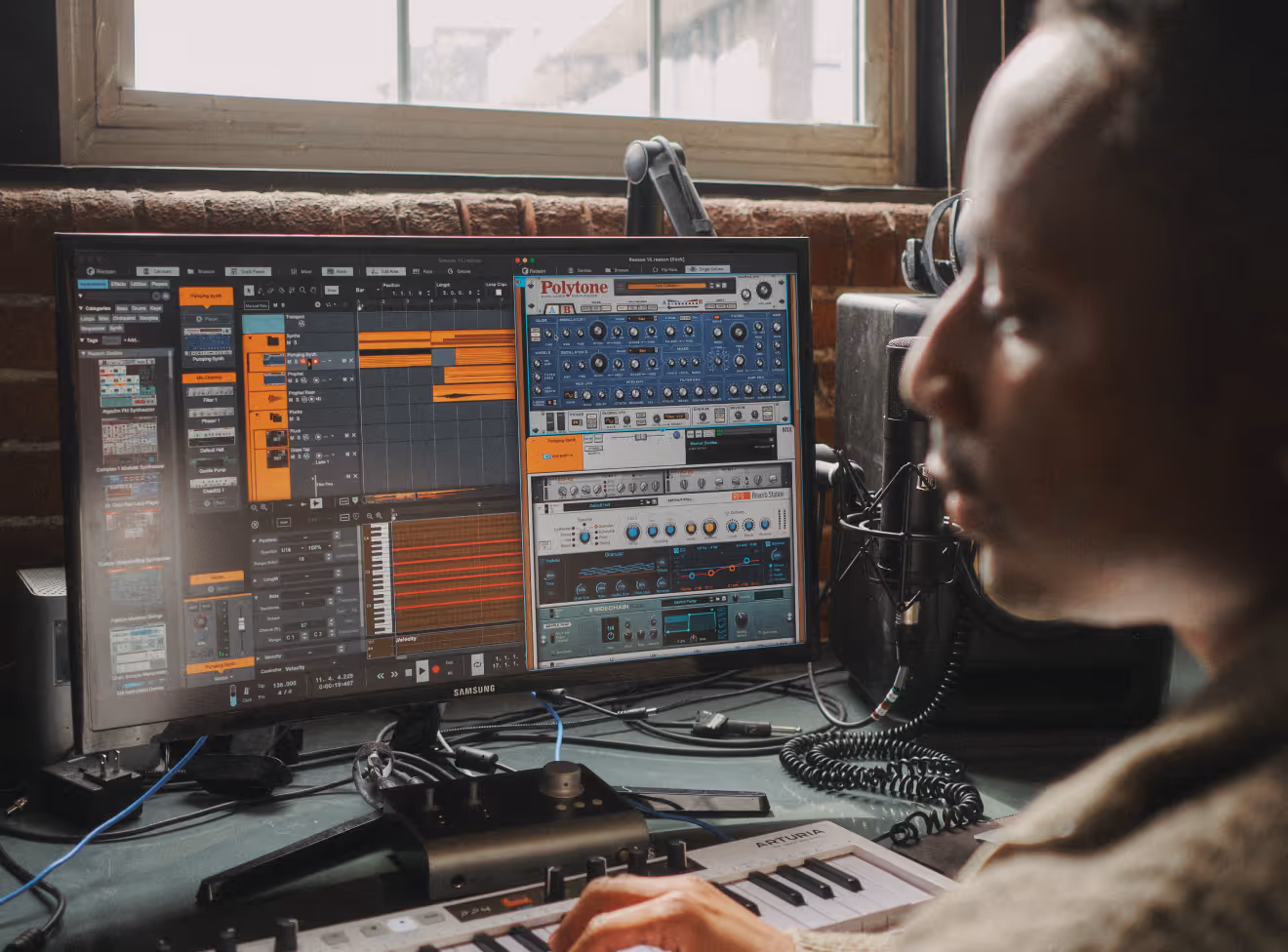 Person using a MIDI keyboard and Reason DAW on a monitor in a studio.