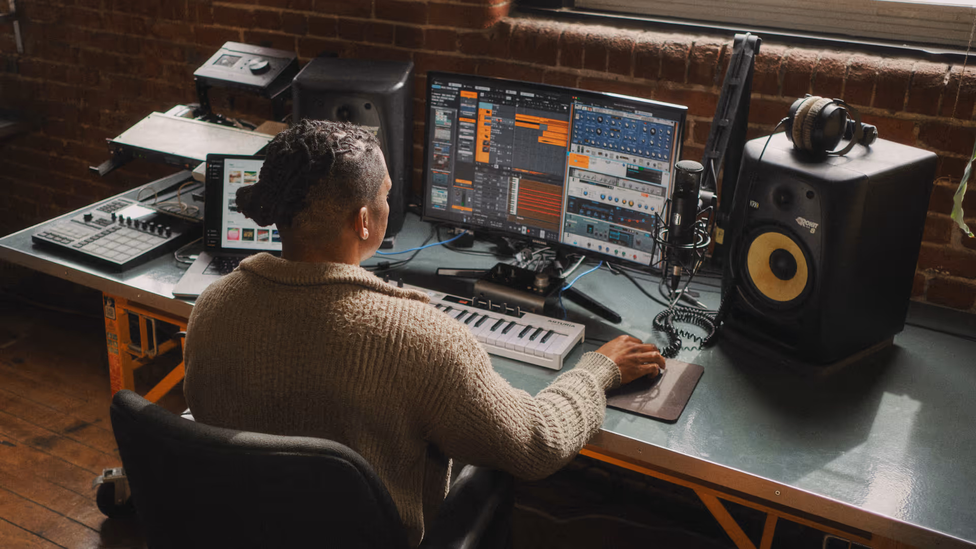 Person working on music production with dual monitors, keyboard, microphone, and speakers in a studio.