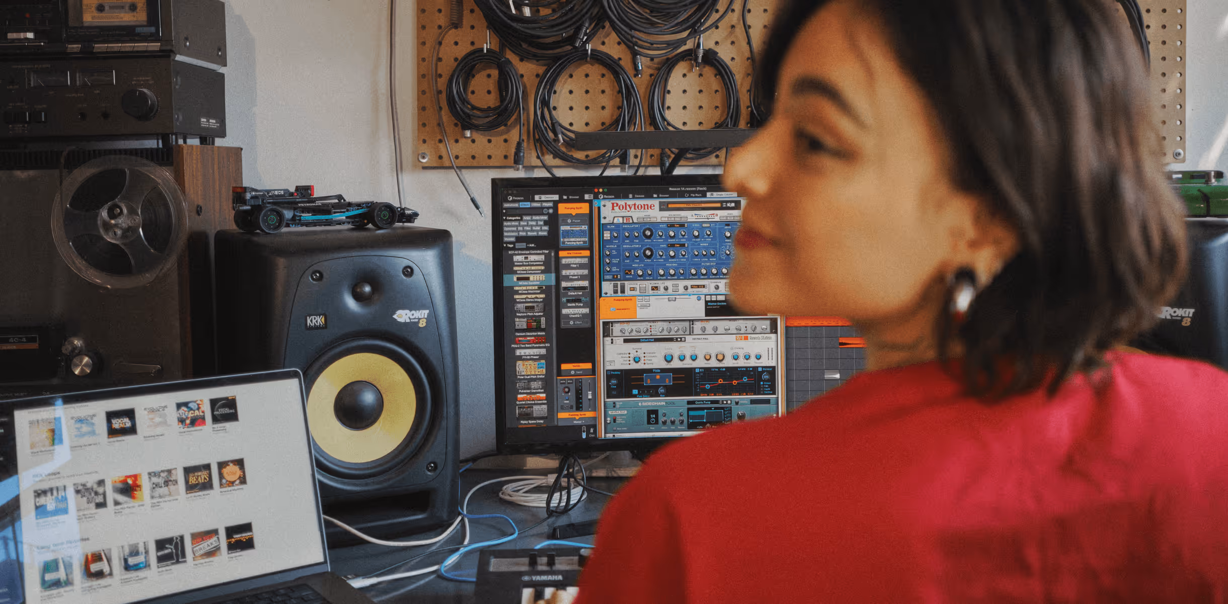 Person working at a music production setup with monitors, speakers, cables, and a laptop with audio files.