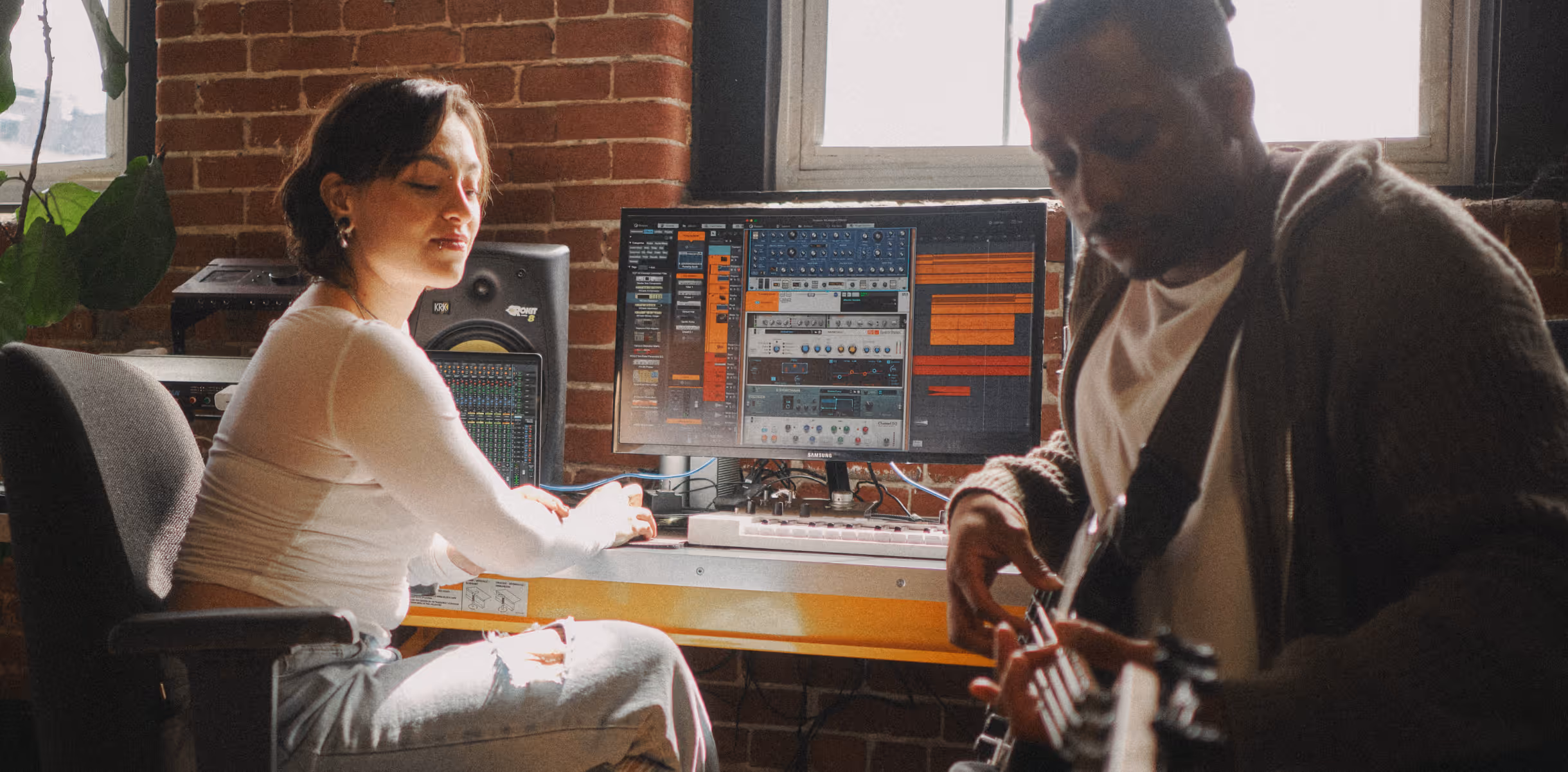Woman working on music production at a computer while a man plays electric guitar in a studio.