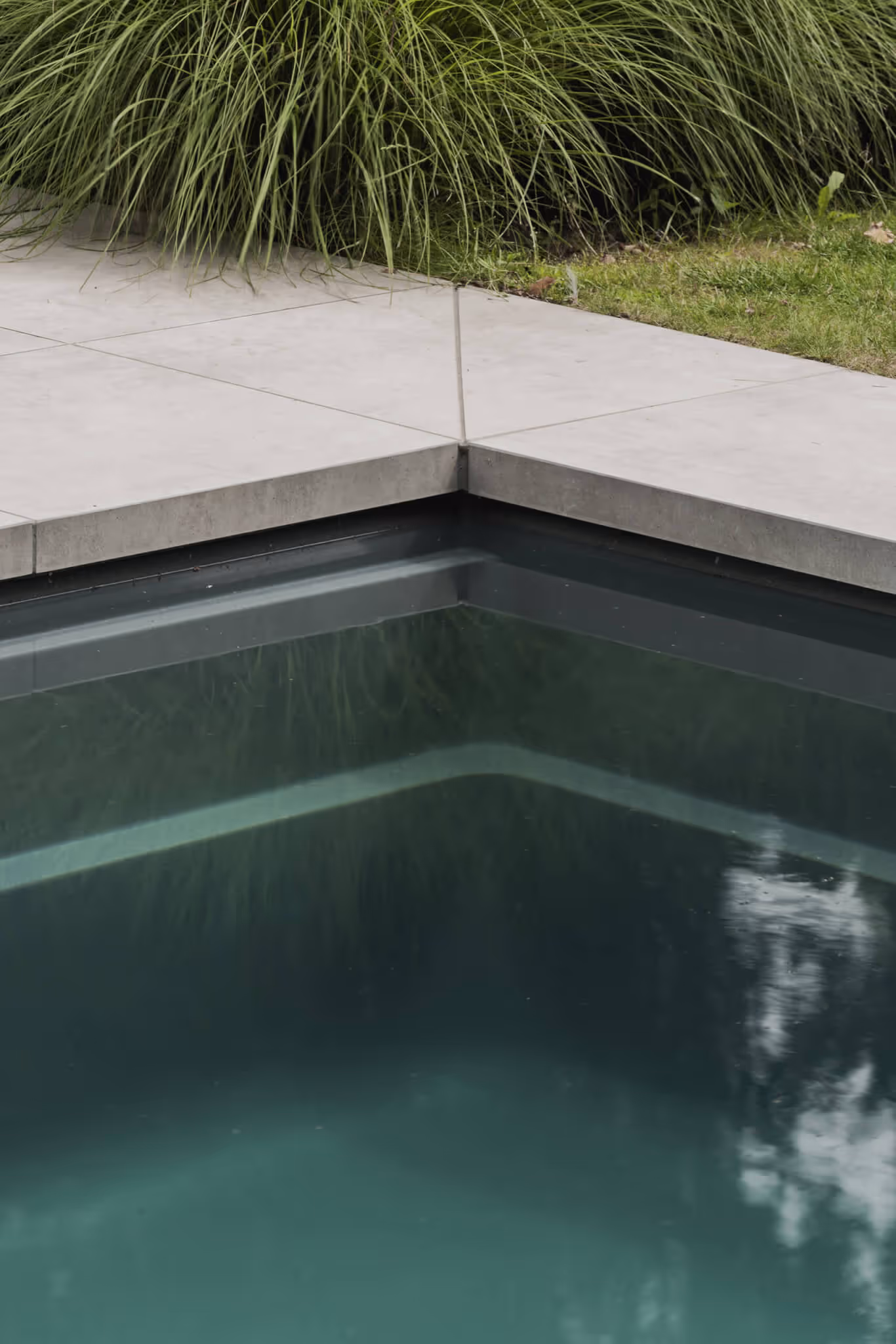 Corner of a modern swimming pool with gray tiles and green grass in the background.
