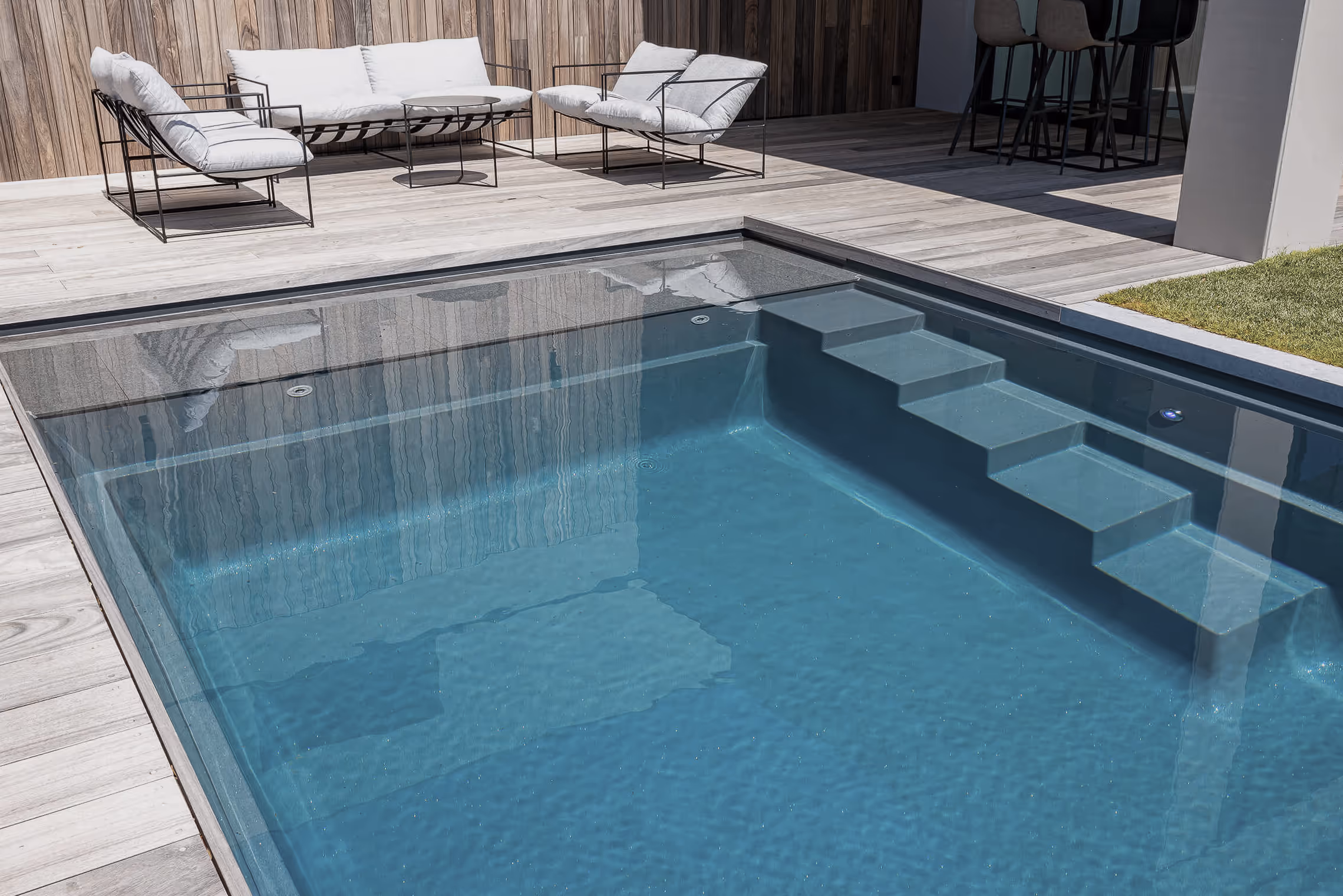 Modern clear pool with submerged blue steps beside wood deck and white cushioned chairs.