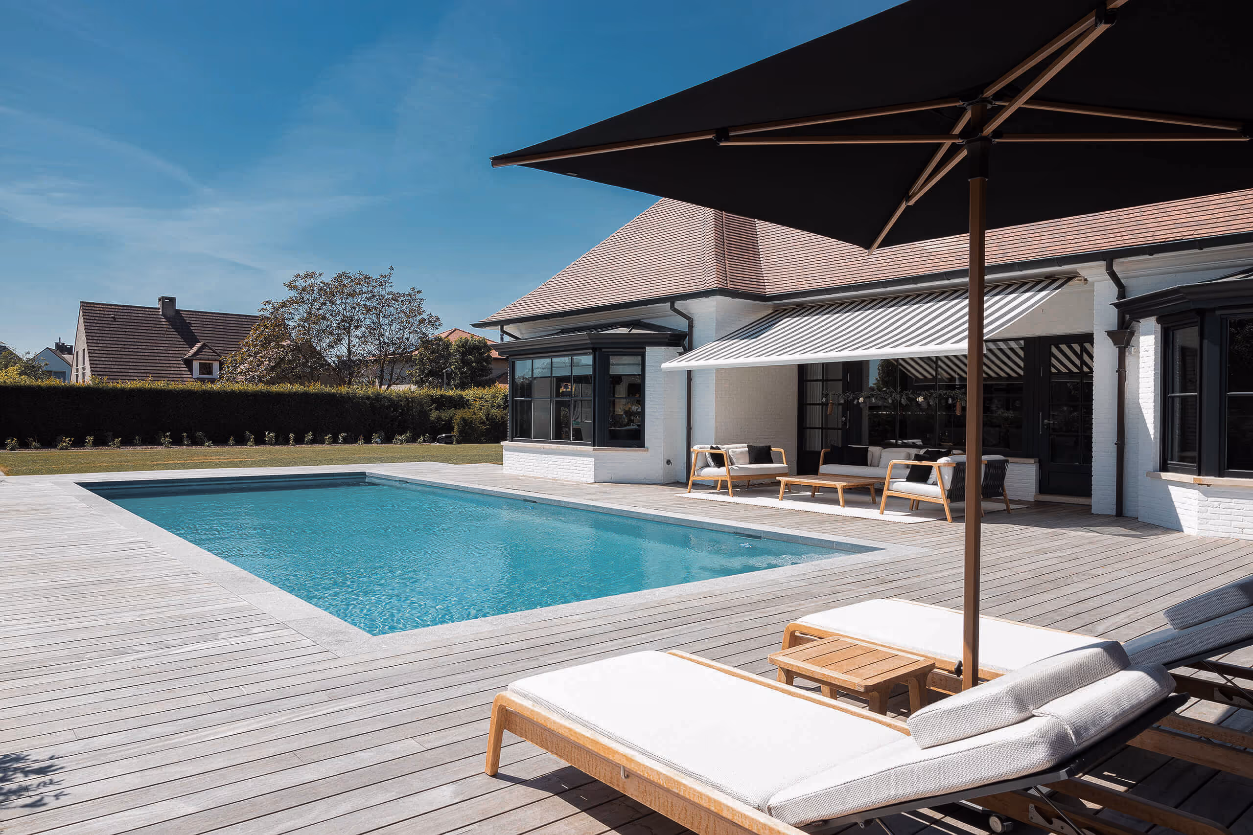 Modern backyard with a rectangular swimming pool, wooden deck, sun loungers, and outdoor seating under an awning.