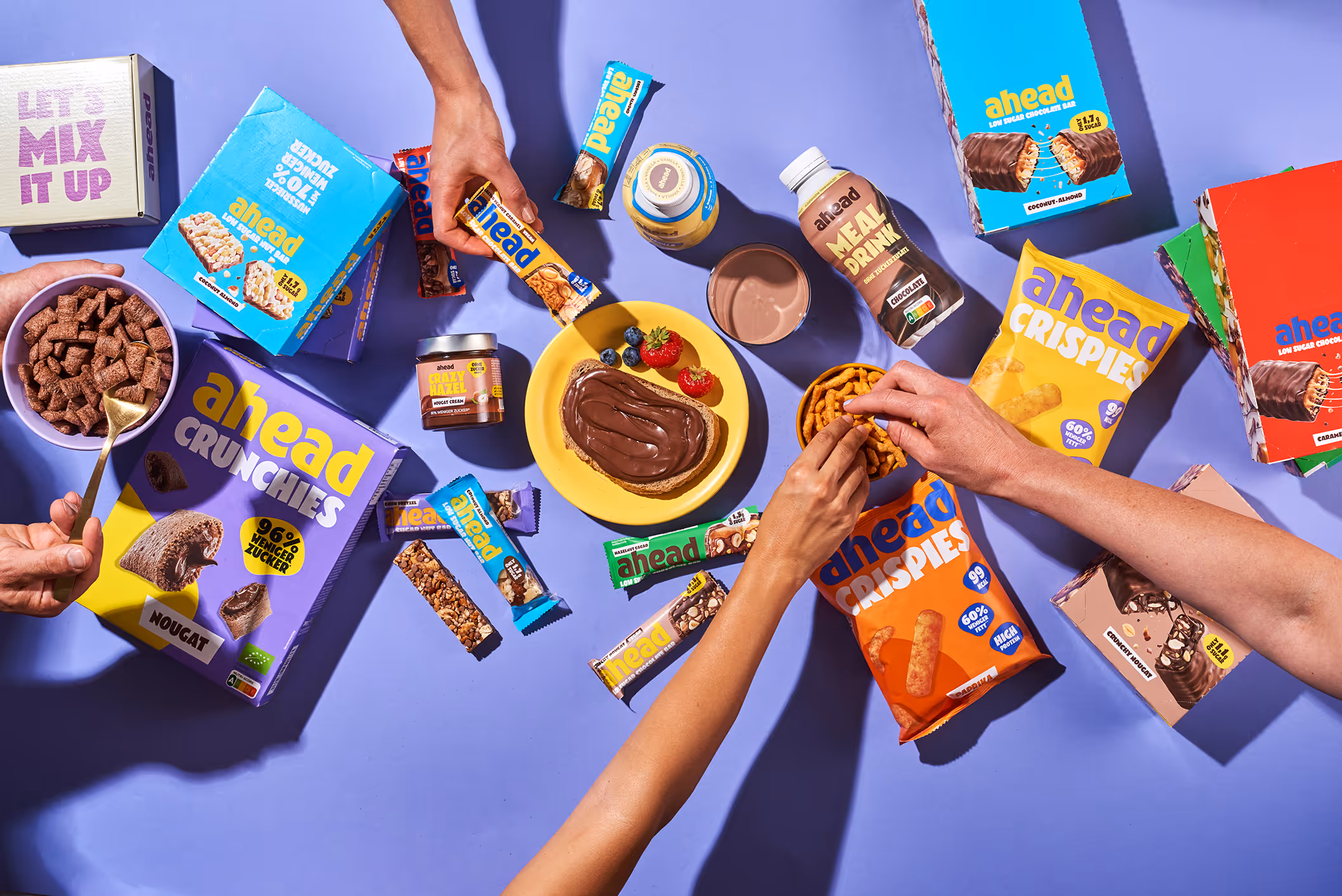 Hands reaching for Ahead brand snacks, bars, cereal, spread, and a meal drink on purple background.