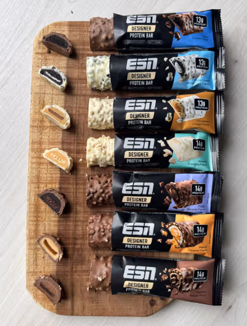Seven ESN Designer Protein Bars in different flavors with cross-section pieces on a wooden board.