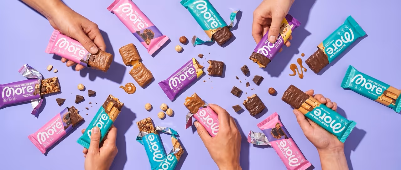 Hands holding and breaking colorful packaged protein bars with chocolate and nuts on lavender background.