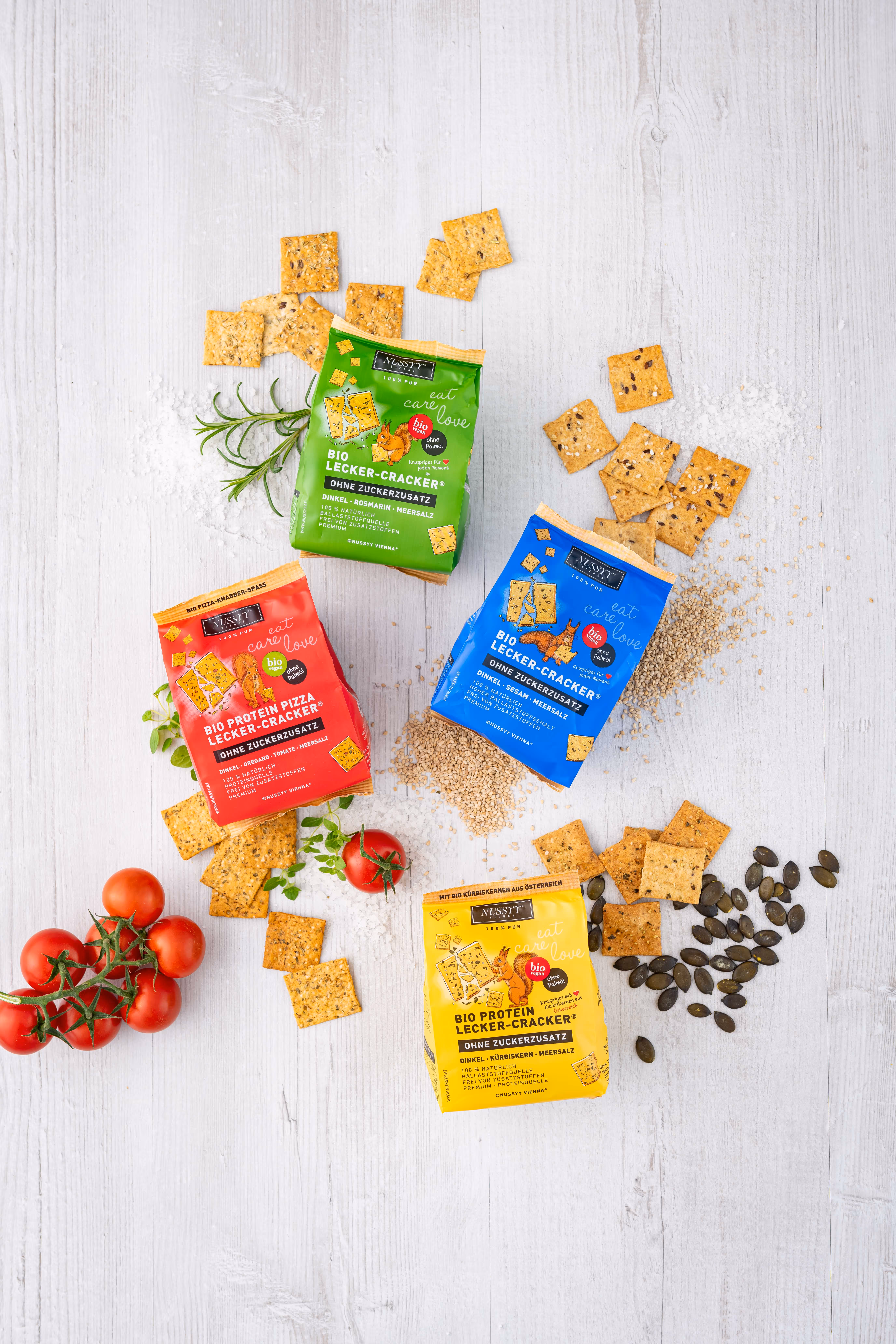 Four colorful packets of organic crackers with scattered crackers, seeds, herbs, and tomatoes on white wood.