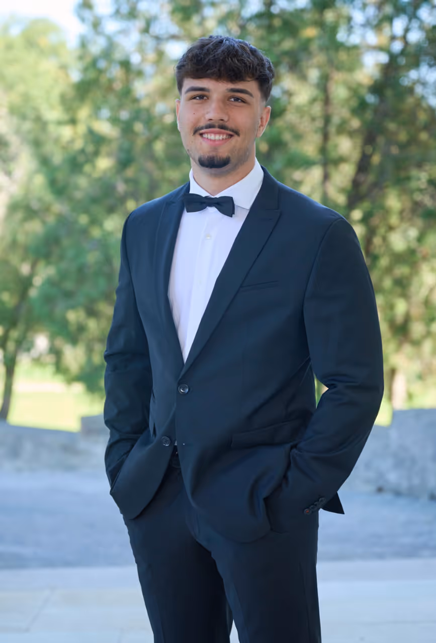 Smiling young man in a black tuxedo with a bow tie standing outdoors with green trees in background.
