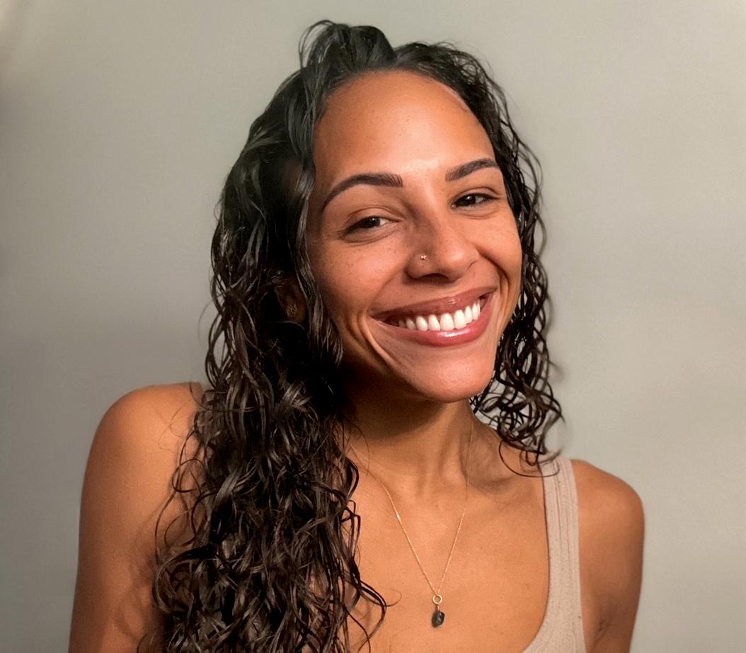 Smiling woman with long curly hair wearing a beige tank top and a necklace with a small pendant.