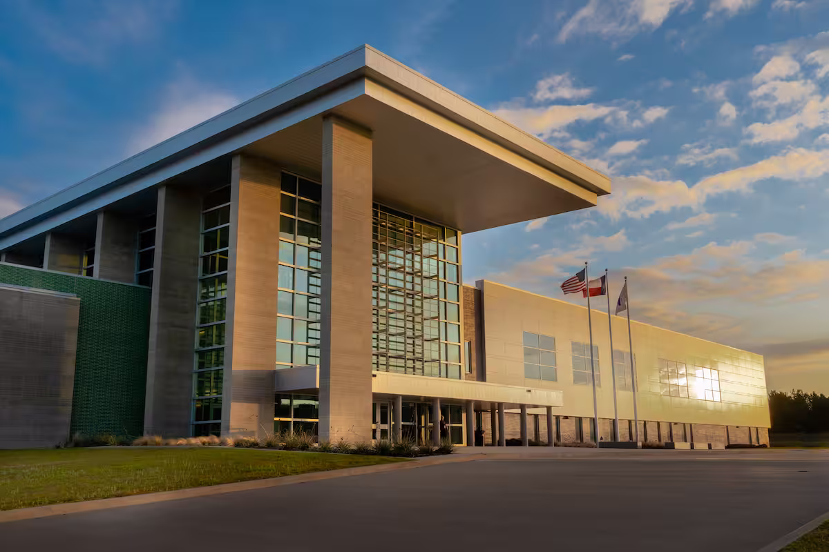 Modern building with large glass windows and three flagpoles under a cloudy sky at sunrise.