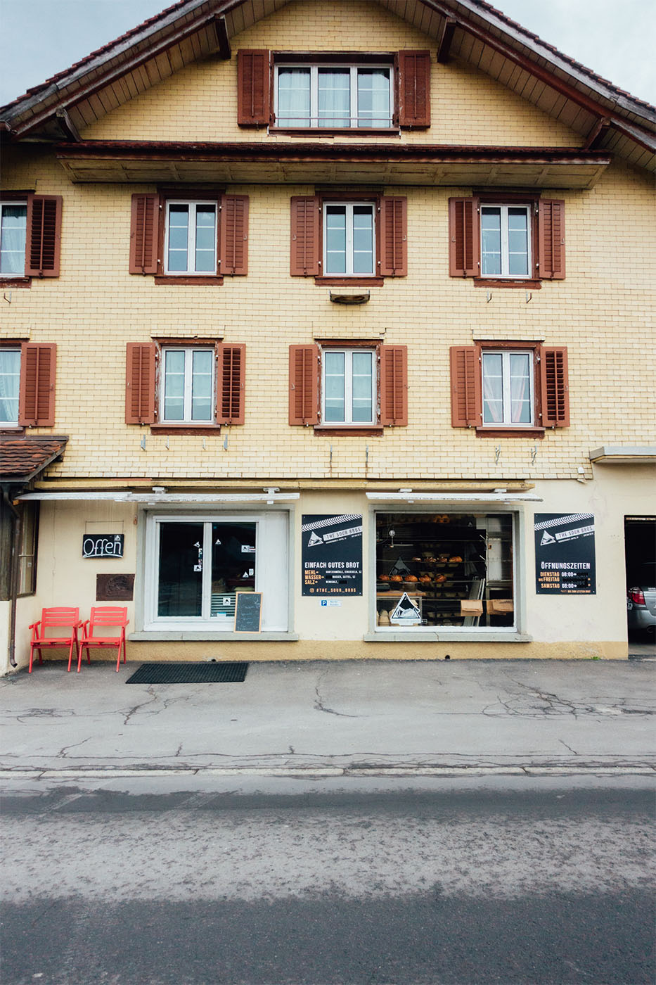 Fassade eines Gebäudes mit sechs Fenstern und einem Laden mit Backwaren, zwei roten Stühlen links und Schildern mit Öffnungszeiten.