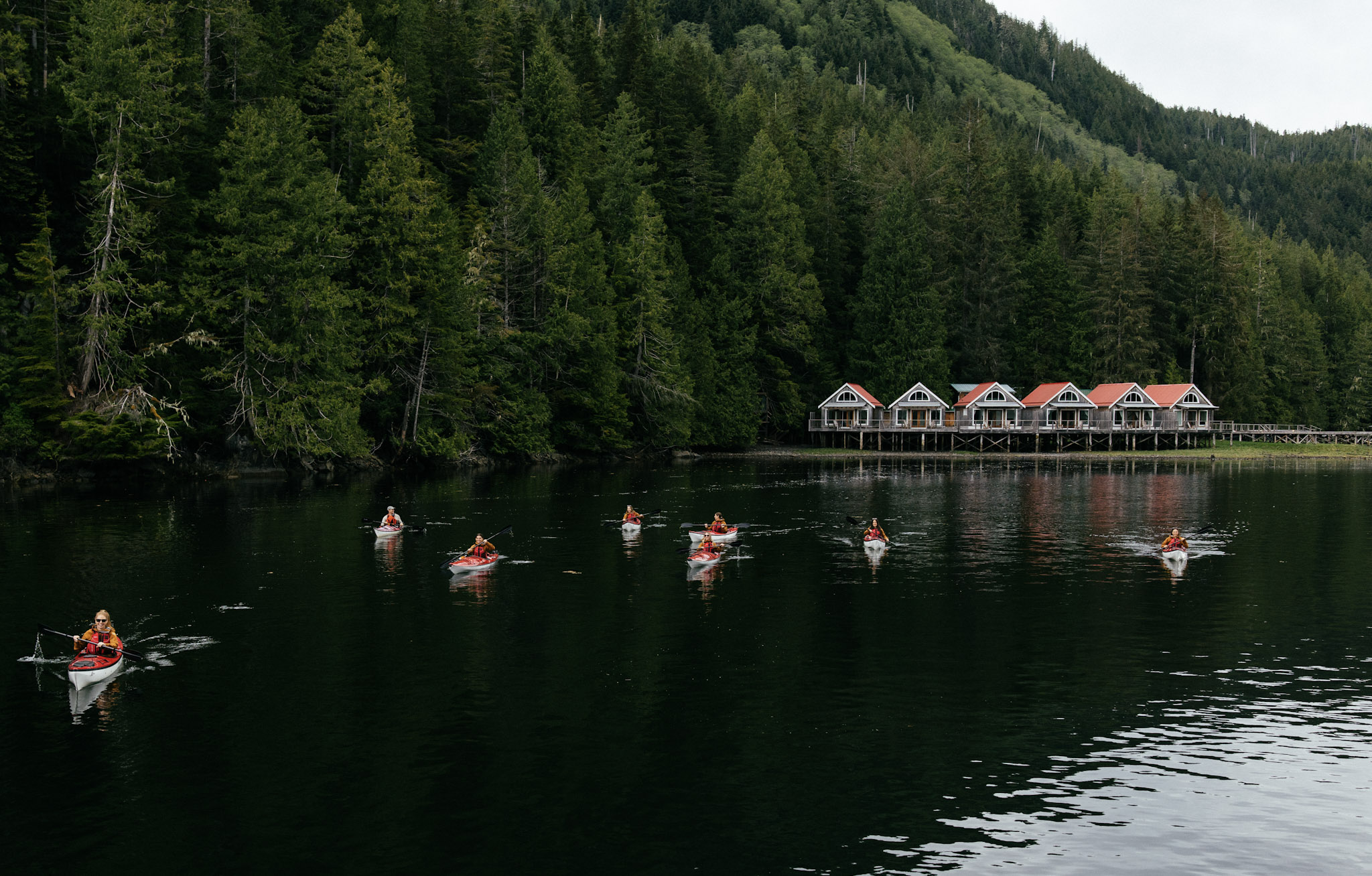 Kayakers paddle past Nimmo Bay's red-roofed waterfront cabins, backed by dense Great Bear Rainforest hillside