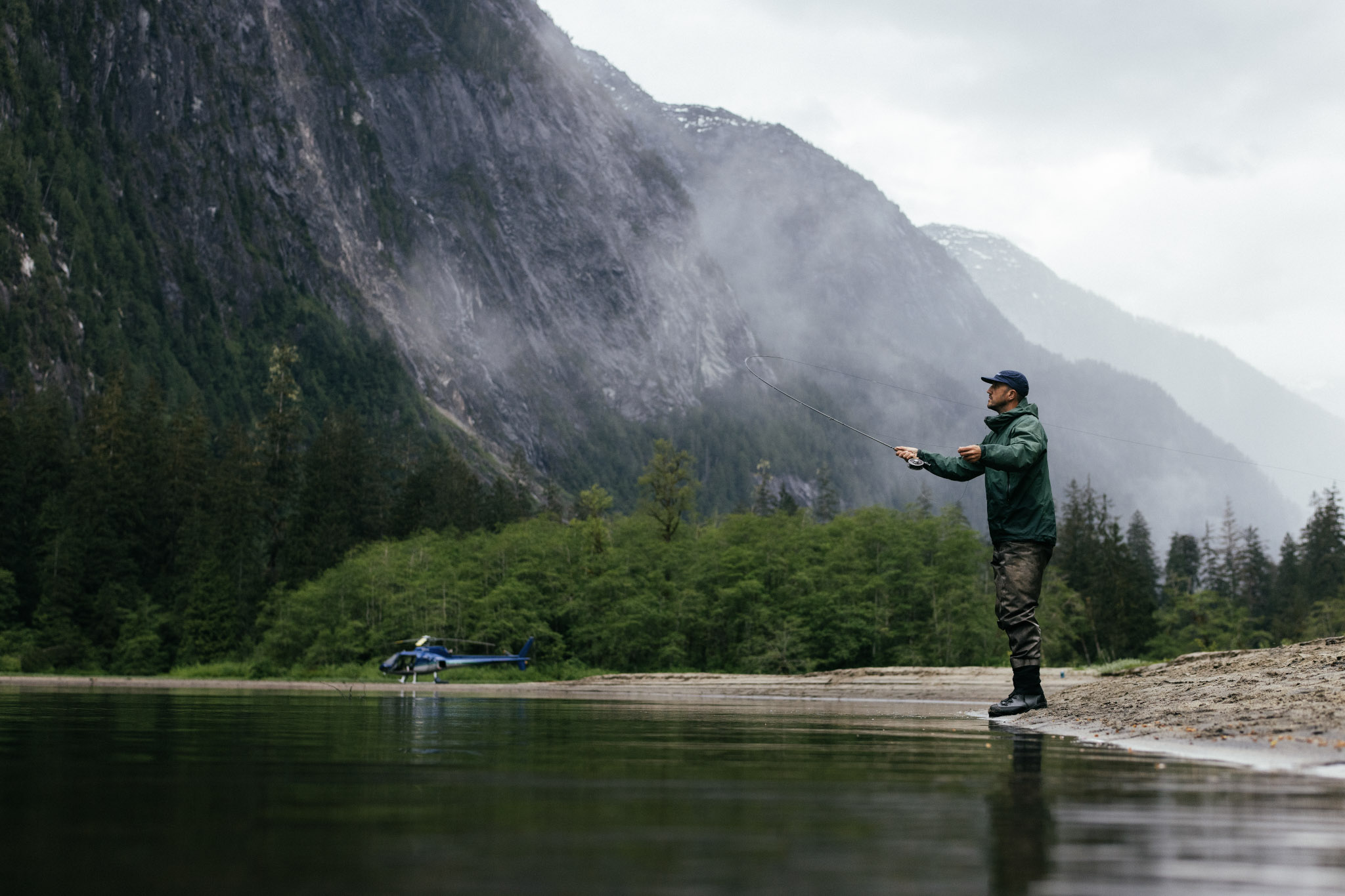 Man fly-fishing. An angler casts on a remote river below a misted mountain wall, the helicopter parked on the gravel bar