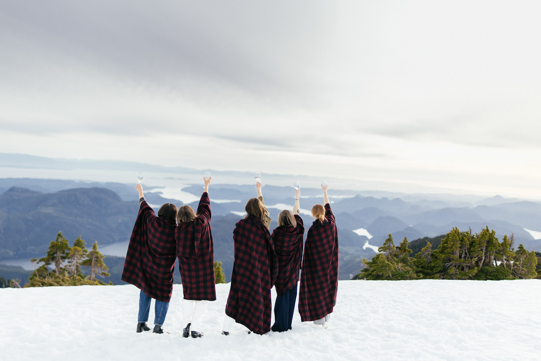Guests wrapped in plaid blankets raise a toast on a snowy ridge above the coastal inlets at Nimmo Bay
