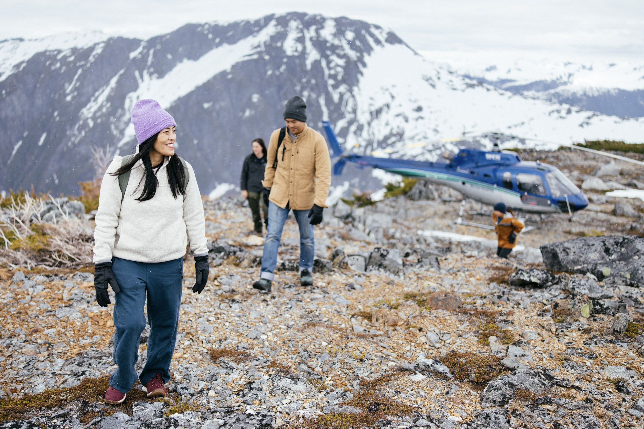 	Guests walk across rocky alpine terrain beside a parked helicopter on a Nimmo Bay heli-excursion