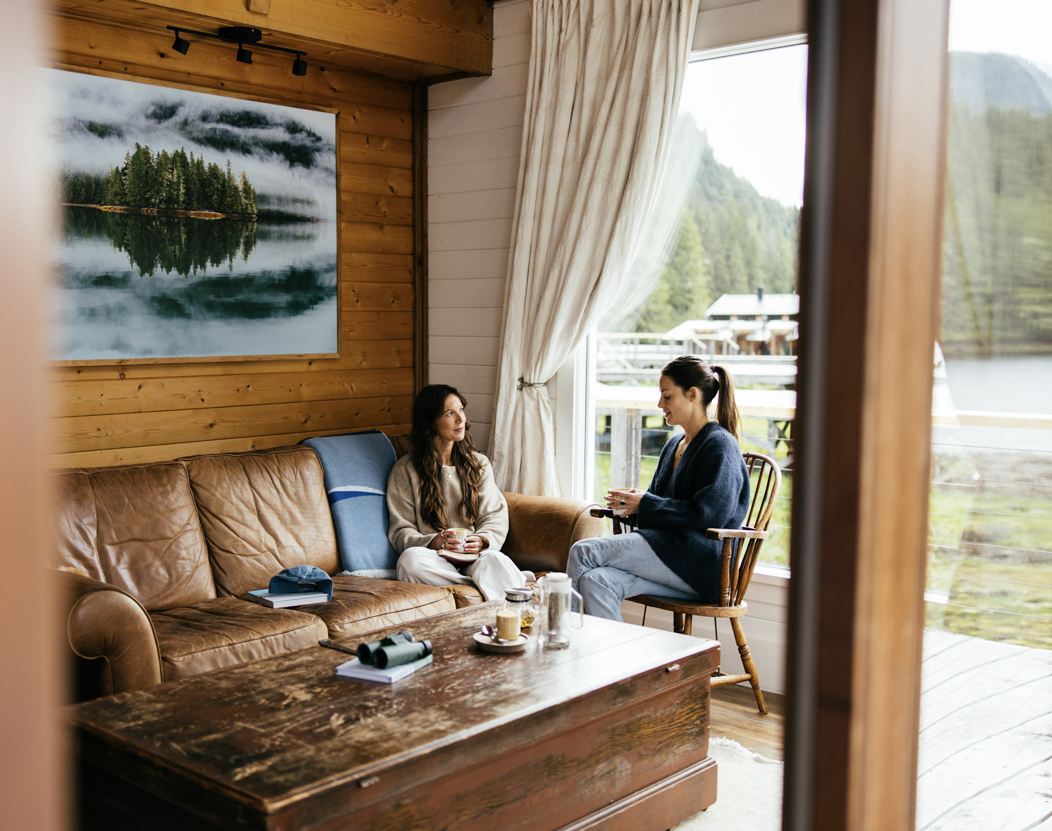 Two guests talk in the lounge of a waterfront cabin at Nimmo Bay, wood-panelled with windows onto the bay